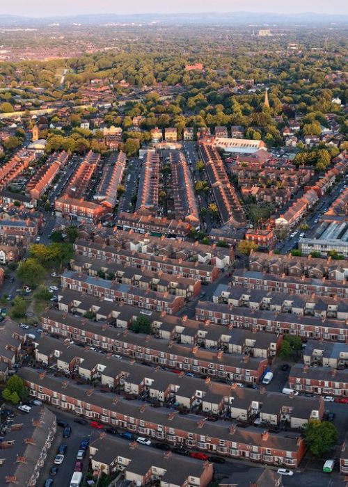 A wide panoramic aerial shot capturing rows of red brick terraced housing across Old Trafford in Greater Manchester, stretching towards leafy suburbs bathed in warm sunset light.